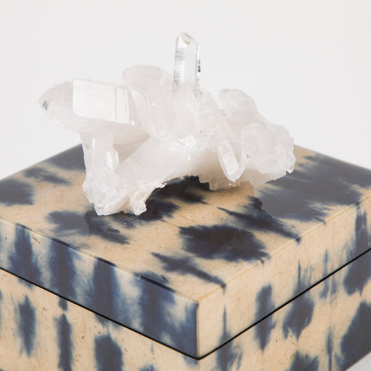 A square box with a geometric pattern, adorned with a blue and black ikat design, and topped with a white quartz crystal.